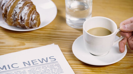 cropped view of man taking coffee with milk near newspaper and croissant.
