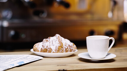 cup of coffee near croissant on plate and newspaper on table.
