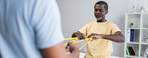 african american man working out with elastics together with blurred rehabilitologist, banner