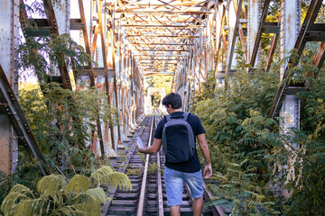 Man talking on a cell phone on a bridge on the train tracks over a river with a backpack. Mention of trips, trails, exploration of new places, technology in isolated places.