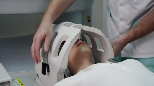 Tracking shot of a young woman lying on a table while a doctor removes a coil on her head at the end of an MRI brain scan.