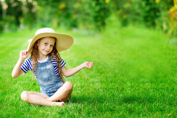 funny baby girl in summer on the lawn with a big straw hat on the green grass having fun and rejoicing, space for text