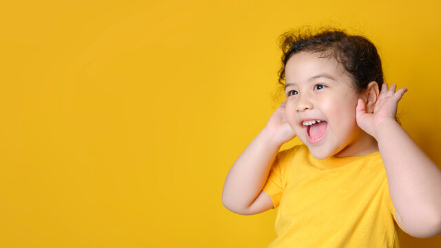Little Asian girl holds her hand near her ear and listenings. Exciting face on Asian child girl. Exciting face on happy asian girl wear yellow shirt and listening to curious good news.