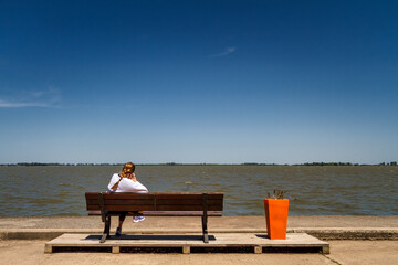 Woman sitting on a bench calls by cell phone in front of the Chascomus lagoon, Argentina
