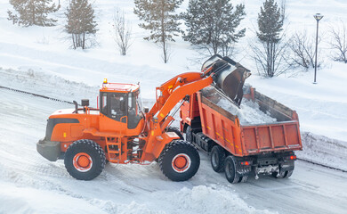 Big orange tractor cleans up snow from the road and loads it into the truck. Cleaning and cleaning...