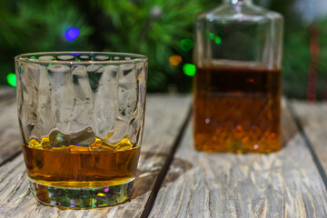 Photo of a glass of whiskey on an old textured wooden table, against the background of a Christmas tree and bottle with alcohol in defocus