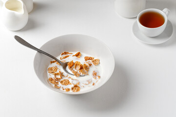 Bowl of wholebread cornflakes with milk on a white table with a cup of tea. Breakfast time. White photography.