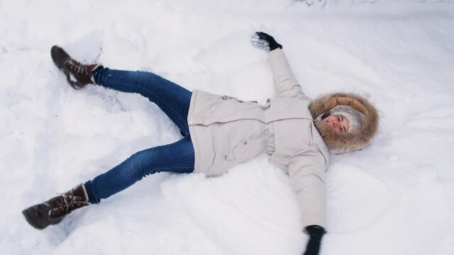 A woman in a white jacket lies in the snow, raises her hands up and down, as if flapping her wings and making an angel
