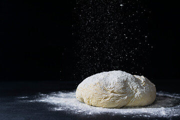 Raw baking dough lies on a dark table. Flour is poured on top. Space for text. Black background. Flour is scattered on the table