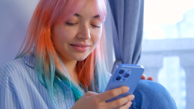 Young woman with dyed hair browsing social media news feed in cellphone. Cute girl with colored hair using mobile app on modern smartphone. Stock video of pretty white female with rainbow hairstyle
