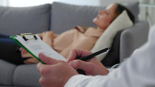 Close up man psychotherapist taking notes on clipboard and listen patient at psychotherapy session. Unfocused adult woman with mental health problem after covid-19 lying on couch talking to therapist