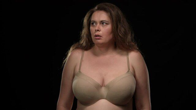 Medium shot Caucasian plus size woman in bra looking around with scared facial expression covering face with hands. Portrait of frightened lady in underwear panicking hiding at black background