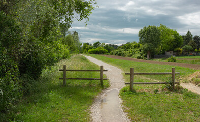 Countryside landscape. White gravel path along the fields with two wooden fences. Puddles on the trail. Cloudy dark sky.