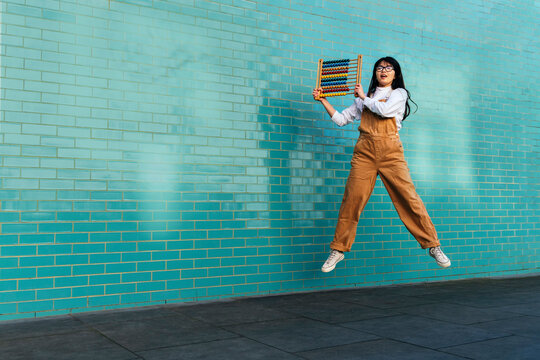 Young woman jumping with abacus in front of turquoise brick wall