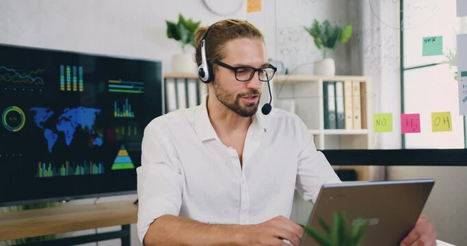 Millennial caucasian male sit at workplace in headset looking at laptop during talking to customer using remote web chat, online video conference application.