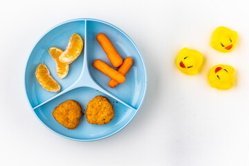 Child food plate divided into three parts filled with chicken nuggets, tangerine pieces and mini carrots placed on white background with rubber duckies on it