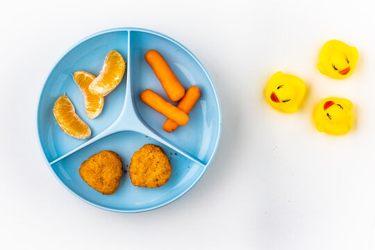 Child Food Plate Divided Into Three Parts Filled With Chicken Nuggets, Tangerine Pieces And Mini Carrots Placed On White Background With Rubber Duckies On It