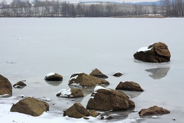 Drained pond and melting snow in winter time. Stones  jutting out over the rest of water. Banks of a pond with trees and hilly landscape  at the background.
