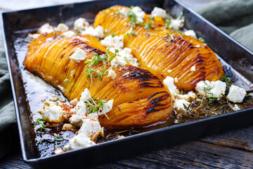 Traditional fried hasselback butternut squash pumpkin roast with herbs and feta sheep cheese served as close-up in a rustic metal tray on a wooden board - obrazy, fototapety, plakaty