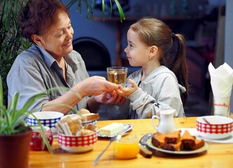 A little granddaughter of 6 years old is serving tea to her grandmother, who is sitting at the table in the kitchen.