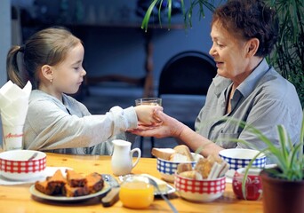 A little granddaughter of 6 years old is serving tea to her grandmother, who is sitting at the table in the kitchen.
