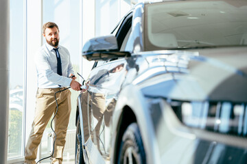 Handsome business man holding charging cable for electric car , looking happy waiting electric car to charge. Caucasian male stands near electric auto in dealership.