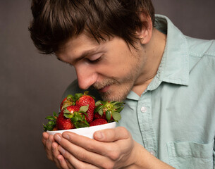 Young handsome tall slim white man with brown hair cherishing strawberries in light blue shirt on...
