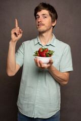 Young handsome tall slim white man with brown hair holding strawberries pointing up in light blue shirt on grey background