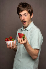 Young handsome tall slim white man with brown hair shocked by a strawberry in light blue shirt on grey background