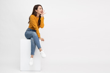 Portrait of young Asian woman sitting on white box and keeps hand near mouth and whispers secret...