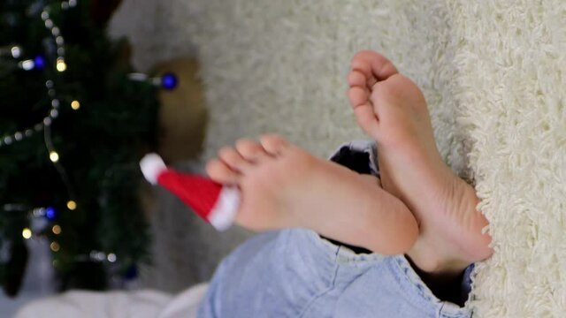 selective focus on child lying on floor on back, legs crossed, santa claus hat on toes against background of blurry stains of garlands, christmas concept