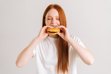 Close-up portrait of pretty young woman with closed eyes enjoying bite of appetizing delicious hamburger on white isolated background in studio. Closeup front view of female eating tasty burger .