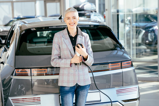 Business woman with the charging cable near the electic vehice, hybrid cars. Positive female salesperson holding charging cable for electric car, looking happy . Concept of buying electric vehicle.