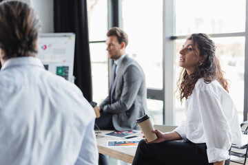 Fototapeta premium businesswoman holding coffee to go while sitting on desk near blurred colleagues.