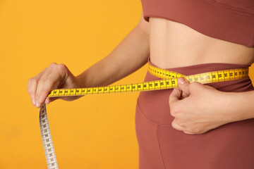 Woman in sportswear measuring waist with tape on yellow background, closeup