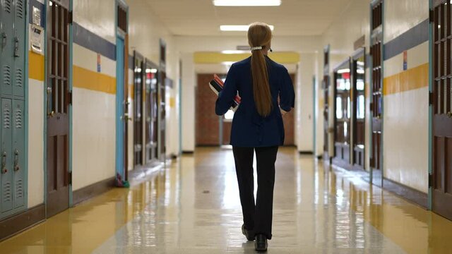 Slow motion rear view of teacher walking down a hallway in an empty school holding books showing emptiness.