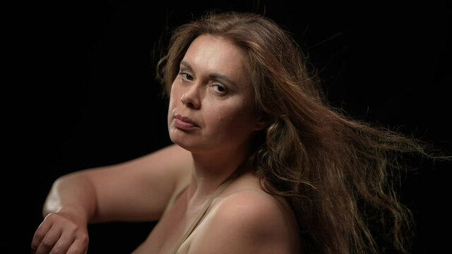 Close-up side view sensual overweight brunette woman with hair blowing by wind posing at black background. Portrait of plus size seductive Caucasian lady looking at camera sitting on chair