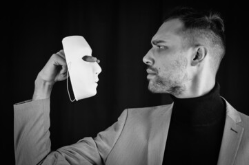 portrait of a young businessman in a gray suit holding and looking intently at a white mask - concept for showing the need to be true and authentic