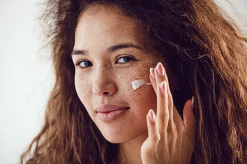 Poster Portrait of woman with curly hair and freckles applying moisturizer to her face. © kanashkin