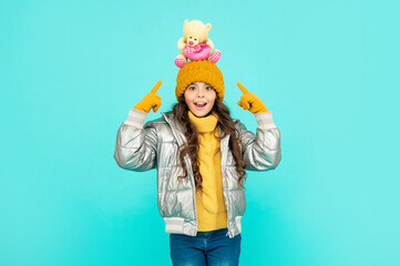 amazed child in hat and puffer jacket point finger on toy on blue background, toy shop