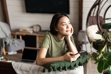 Dreamy asian girl spend time at home, holding smartphone and sitting on couch, smiling while...