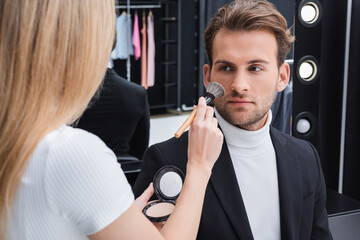 blurred visage artist applying face powder with cosmetic brush on man in photo studio.