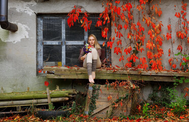 A beautiful young woman is seen relaxing by an old house.
She is surrounded by falling red autumn...