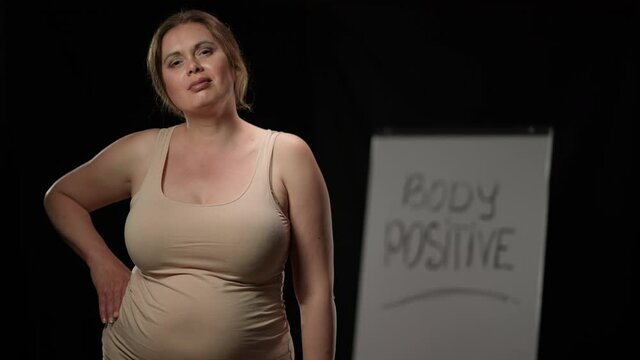 Brunette plus size Caucasian woman gesturing thumb up looking at camera with blurred Body positive on whiteboard at black background. Portrait of confident obese lady posing smiling