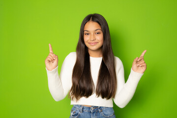Brunette smiling hispanic girl wearing white sweater pointing fingers up over green background.