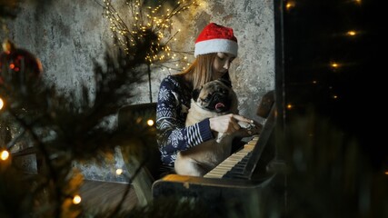 Teenager girl in santa hat playing a christmas song on the piano together with a funny pug dog, new year mood