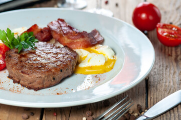 large grilled steak on a white plate with tomatoes fried bacon and poached egg
