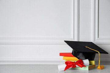 Graduation hat, books and diploma on floor near white wall, space for text