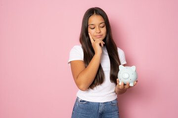 Pensive brunette girl in casual t-shirt holding a piggy bank standing isolated over pink background.