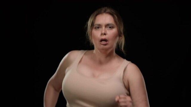 Motivated plus size sportswoman running at black background looking at camera. Portrait of inspired confident Caucasian overweight lady training posing. Sport and self improvement